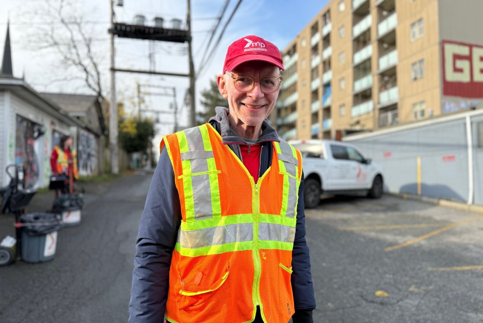 Arwin is wearing a red Mission Possible hat, grey sweater, and an orange and yellow safety vest. He is outside in a sunny alleyway. 