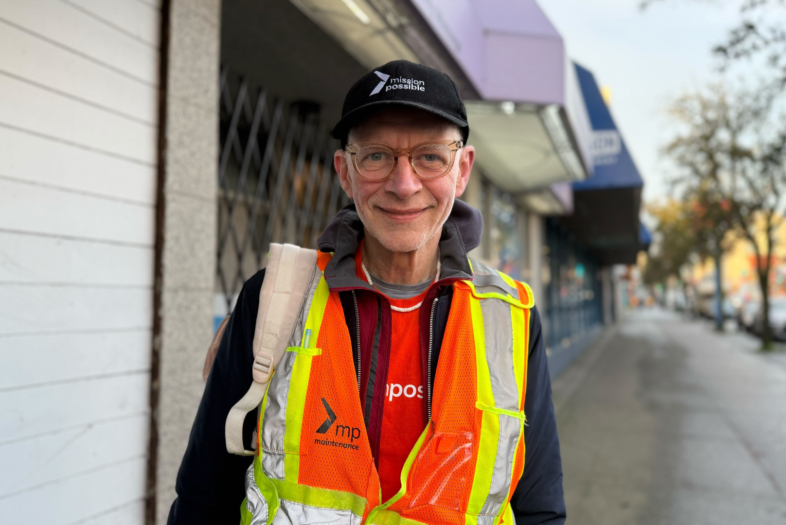 Arwin has glasses and is wearing a black Mission Possible project bridge hat, a black jacket, orange shirt, and an orange and yellow safety vest. 