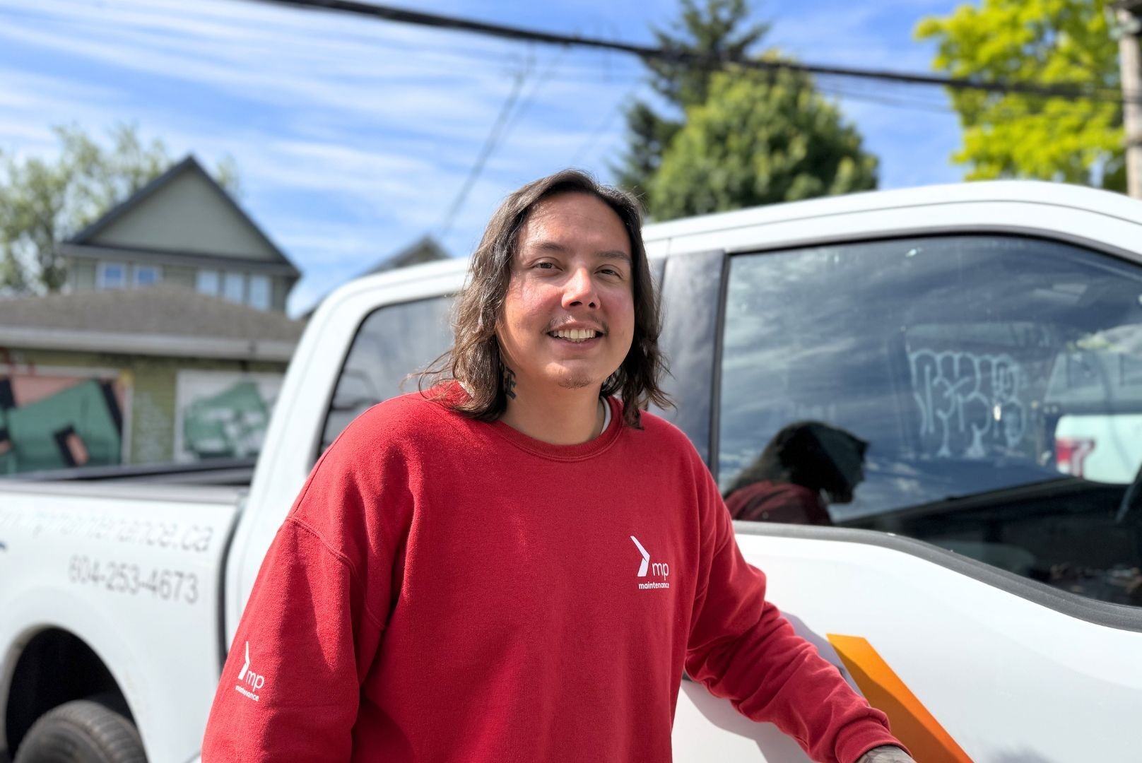 Black is wearing a red Mission Possible sweatshirt and is leaning against a white truck. It is a sunny blue sky day. 