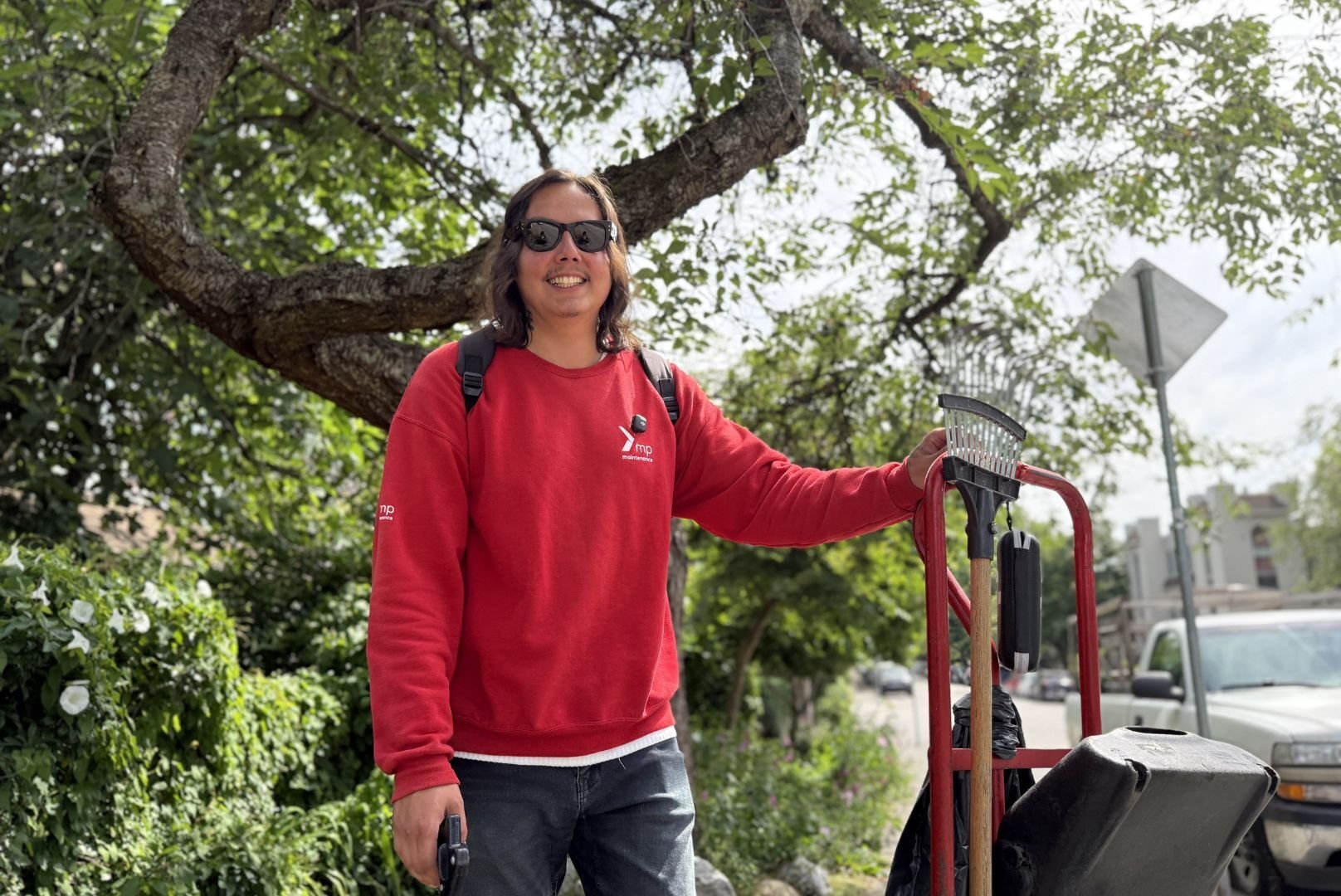 Black is wearing a red Mission Possible sweatshirt and sunglasses. He is standing under a tree smiling next to his cleaning cart. 