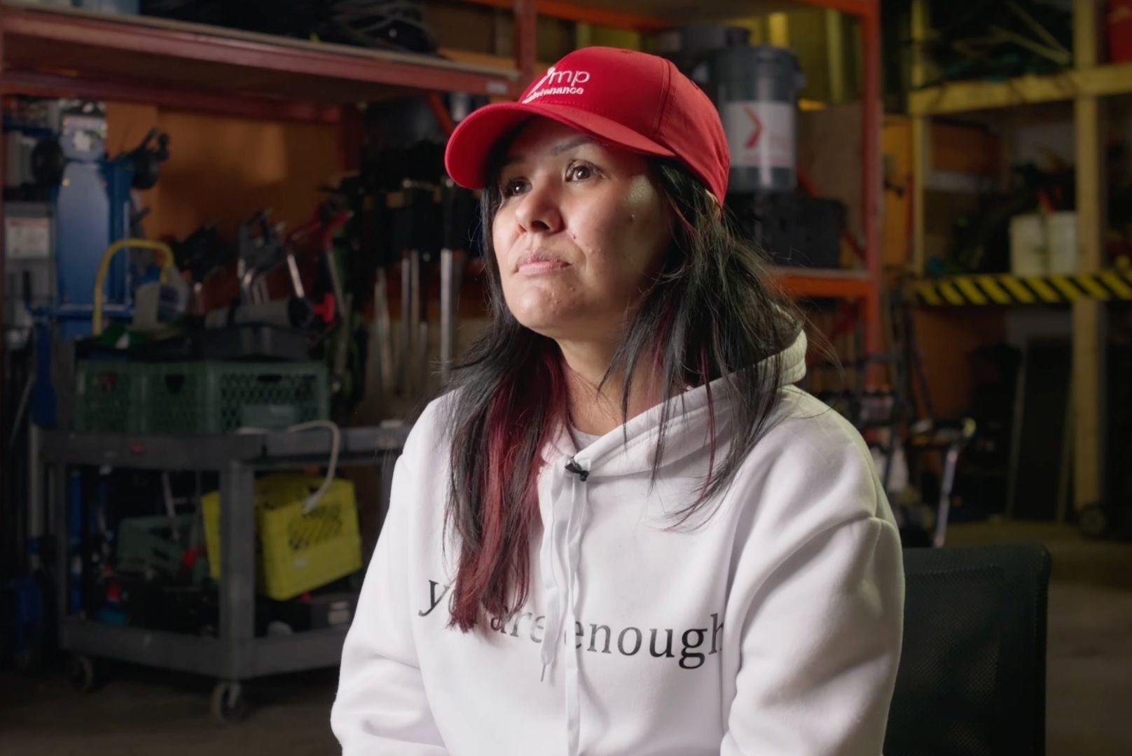 Rochelle is wearing a white hoodie and red MP Maintenance hat. She has long hair that goes past her shoulders. She is in the MP warehouse. 