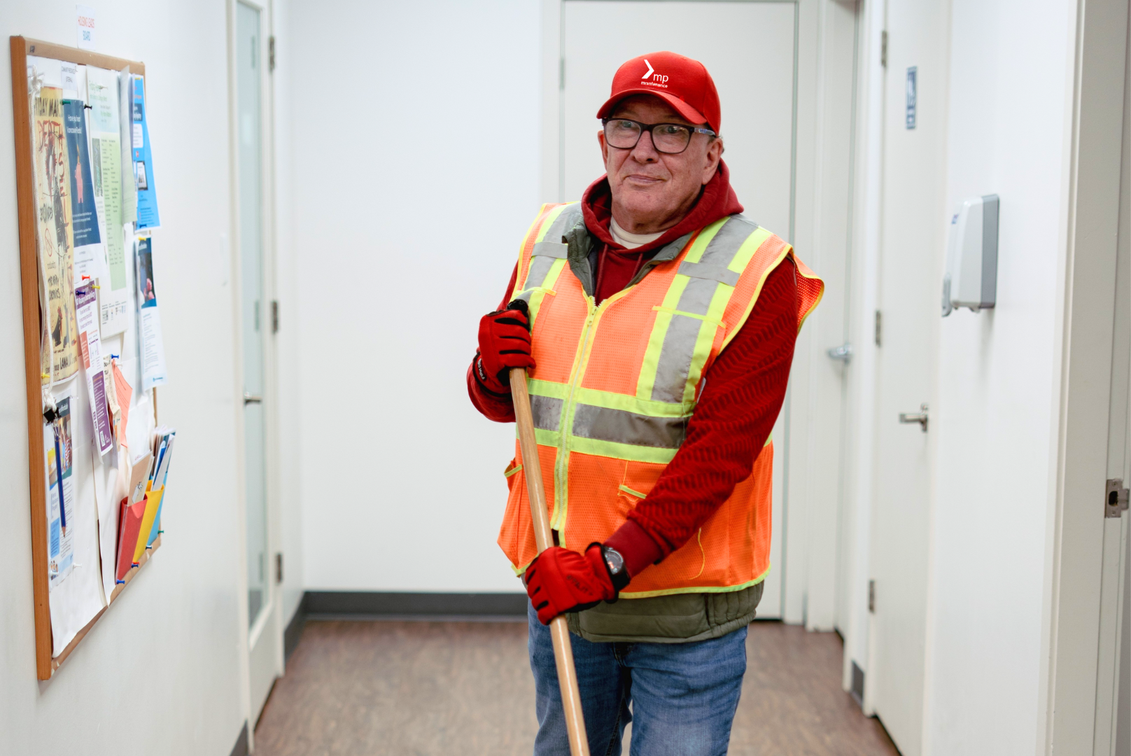 Rodney is wearing a red Mission Possible hat, red hoodie, and an orange and yellow safety vest with jeans. He is in the Mission Possible office doing janitorial services.